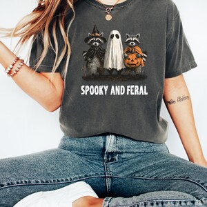 May include: Dark gray t-shirt with the text "SPOOKY AND FERAL". The shirt features a Halloween design with raccoons dressed as a witch and holding a pumpkin, and a ghost. The shirt is made of a soft material.