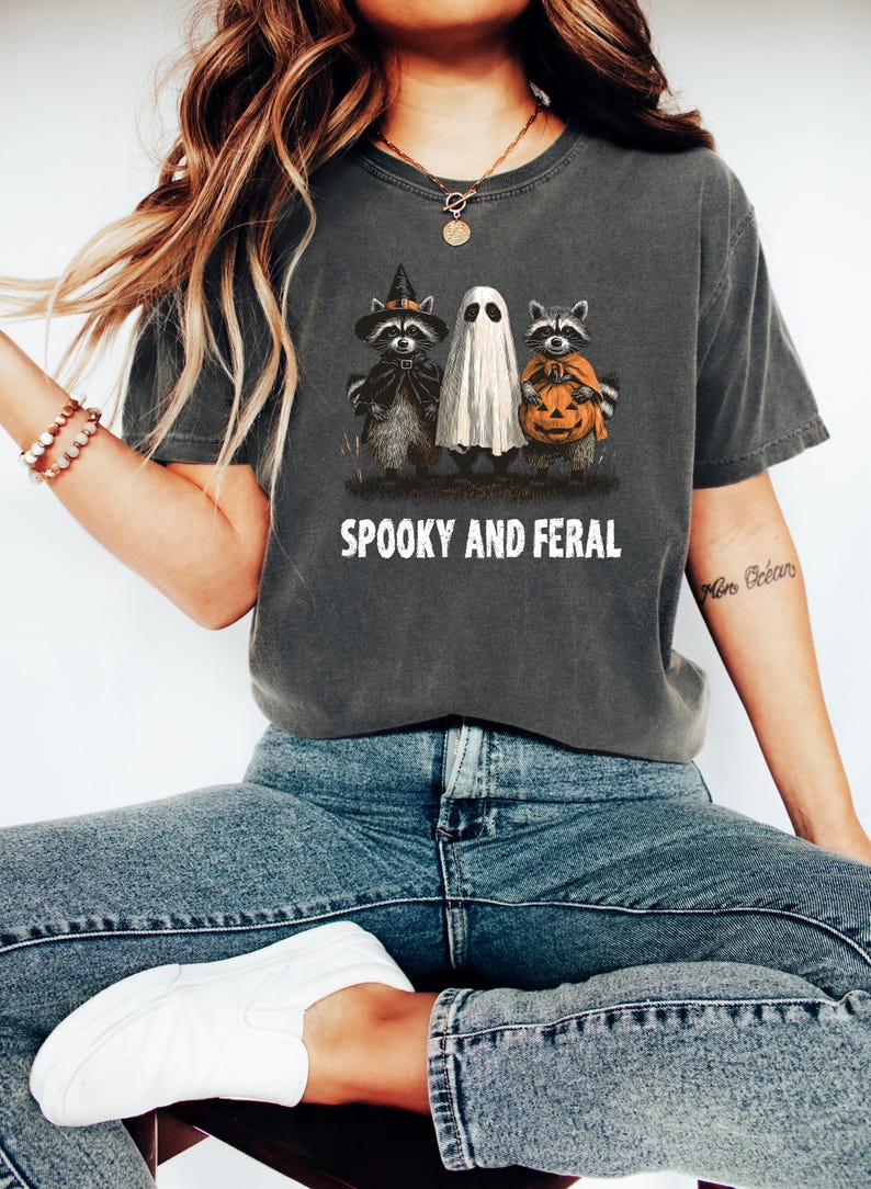 May include: Dark gray t-shirt with the text "SPOOKY AND FERAL". The shirt features a Halloween design with raccoons dressed as a witch and holding a pumpkin, and a ghost. The shirt is made of a soft material.