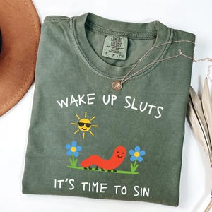 May include: Olive green t-shirt with the text "WAKE UP SLUTS IT'S TIME TO SIN". The graphic includes a smiling sun, a red worm, and blue flowers. A gold necklace is draped across the shirt.