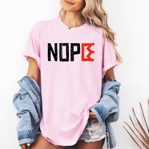 May include: A pale pink t-shirt featuring the word "NOPE" in black, block letters, and a red crown graphic. The shirt has rolled-up sleeves and a relaxed fit.
