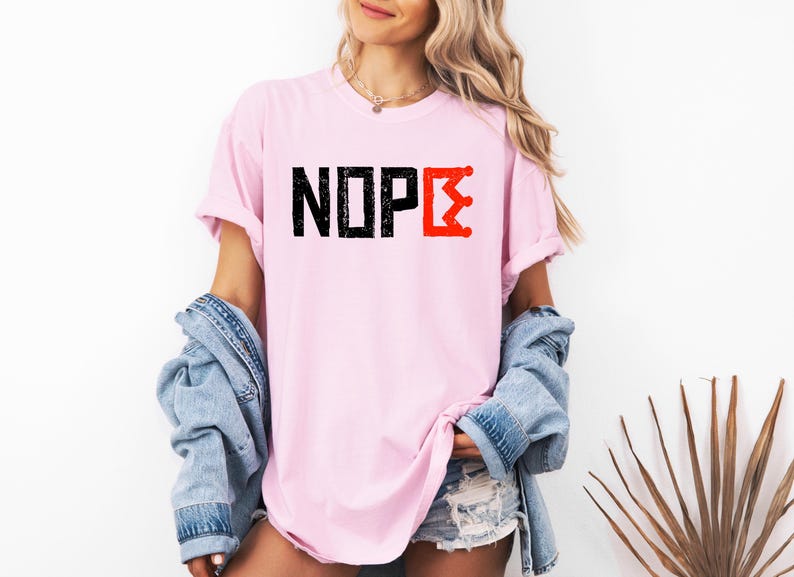 May include: A pale pink t-shirt featuring the word "NOPE" in black, block letters, and a red crown graphic. The shirt has rolled-up sleeves and a relaxed fit.