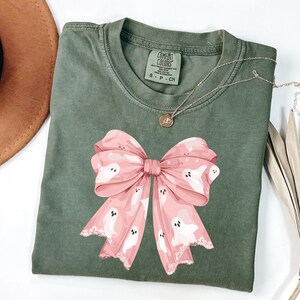 May include: An olive green t-shirt featuring a large pink bow with white ghost illustrations. The bow has a lace-like trim. A gold necklace with a small pendant is on the shirt. The shirt has a Comfort Colors tag.