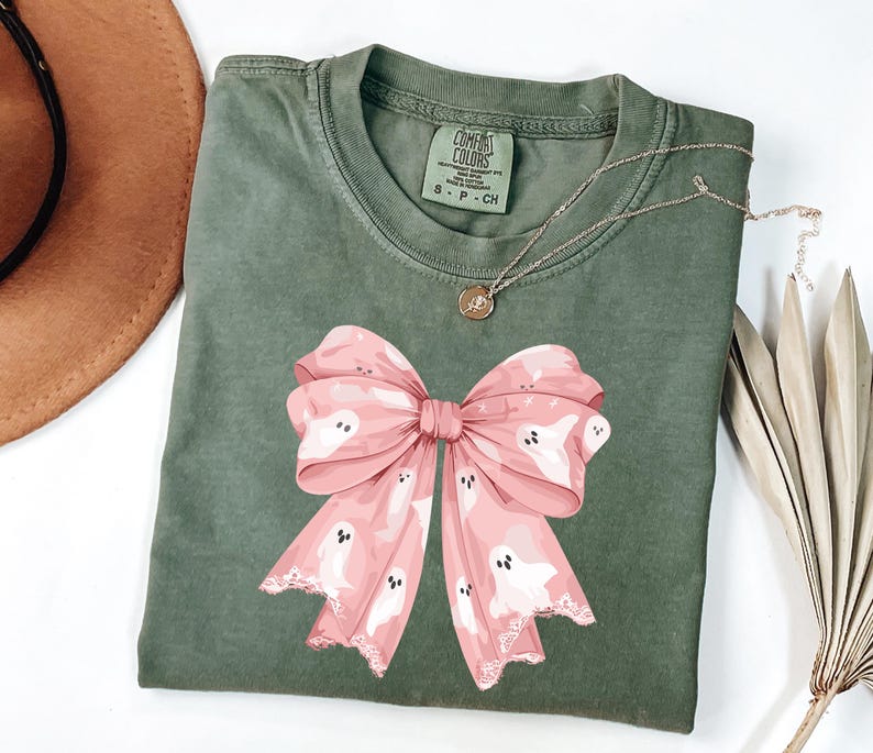 May include: An olive green t-shirt featuring a large pink bow with white ghost illustrations. The bow has a lace-like trim. A gold necklace with a small pendant is on the shirt. The shirt has a Comfort Colors tag.