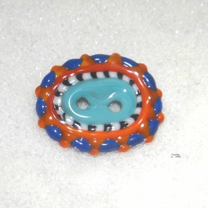May include: A colorful glass button with a turquoise center and a black and white striped ring. The button is surrounded by a ring of orange and blue glass with white dots.