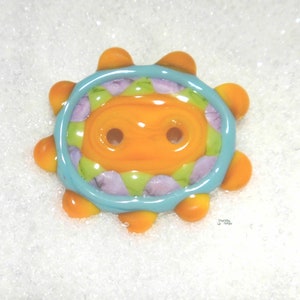 May include: A bright orange button with a blue and green border and purple accents. The button has two holes for sewing.