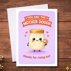 May include: A greeting card with a cartoon jar of dough holding flowers. The card reads "You are the Mother Dough, thanks for rising me!" on a purple background with hearts. The card is on a wooden surface.