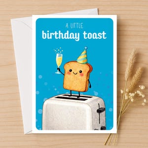 May include: A birthday greeting card with a cartoon illustration of a slice of toast wearing a party hat and holding a champagne glass. The card has the text "A LITTLE birthday toast" on a blue background, with a white envelope.