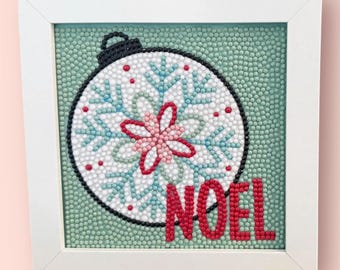 May include: A framed Christmas ornament design with a white frame. The ornament is white with blue snowflake designs and a pink flower in the center. The word "NOEL" is in red at the bottom. The background is a light green.