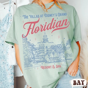 May include: A light green t-shirt with the text "The Villas at Disney's Grand Floridian Resort & Spa" in pink and blue. The design features an illustration of a resort building with palm trees and other architectural details.