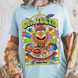 May include: Light blue t-shirt featuring a colorful graphic of Dr. Teeth and The Electric Mayhem from Disney's The Muppets. The design includes the band members and the words "Dr. Teeth" and "The Electric Mayhem World Tour."