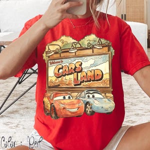 Op de afbeelding: Rood T-shirt met een afbeelding van het Cars Land-bord en Lightning McQueen en Sally Carrera. Het bord leest "Welcome to Cars Land" boven het Cars Land-logo. Het shirt is effen rood van kleur.