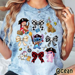 Może przedstawiać: Jasnoniebieska koszulka tie-dye z r&oacute;żnymi postaciami pirat&oacute;w z kresk&oacute;wek i wzorami kokard. Koszulka zawiera wizerunki Stitcha, kaczki i innych postaci w strojach pirackich, a także dekoracyjne kokardy.