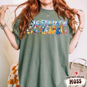 Puede incluir: Una camiseta verde musgo Comfort Colors con un gr&aacute;fico colorido de personajes de Disney, incluyendo Mickey Mouse, Minnie Mouse, Donald Duck y Winnie the Pooh. La palabra "Disneyland" est&aacute; impresa encima de los personajes en letras azules.