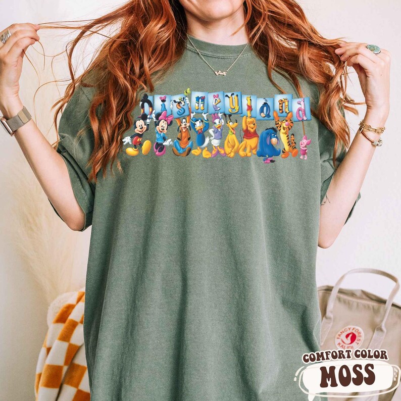 Puede incluir: Una camiseta verde musgo Comfort Colors con un gr&aacute;fico colorido de personajes de Disney, incluyendo Mickey Mouse, Minnie Mouse, Donald Duck y Winnie the Pooh. La palabra "Disneyland" est&aacute; impresa encima de los personajes en letras azules.