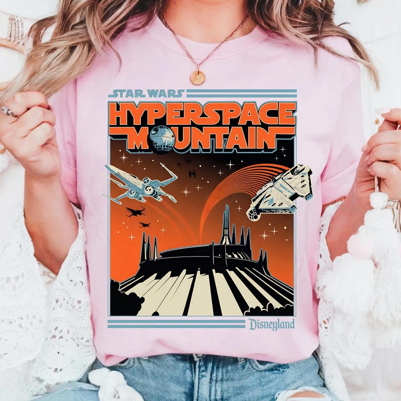 May include: Pink t-shirt featuring a retro-style graphic for Star Wars: Hyperspace Mountain at Disneyland. The design includes the text "Star Wars" and "Hyperspace Mountain" with illustrations of an X-wing, the Millennium Falcon, and the Death Star.