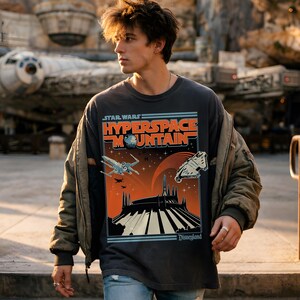 May include: Dark gray Star Wars Hyperspace Mountain t-shirt with a graphic of a spaceship, mountain, and the Disneyland logo. The shirt is worn with a green jacket and blue jeans.