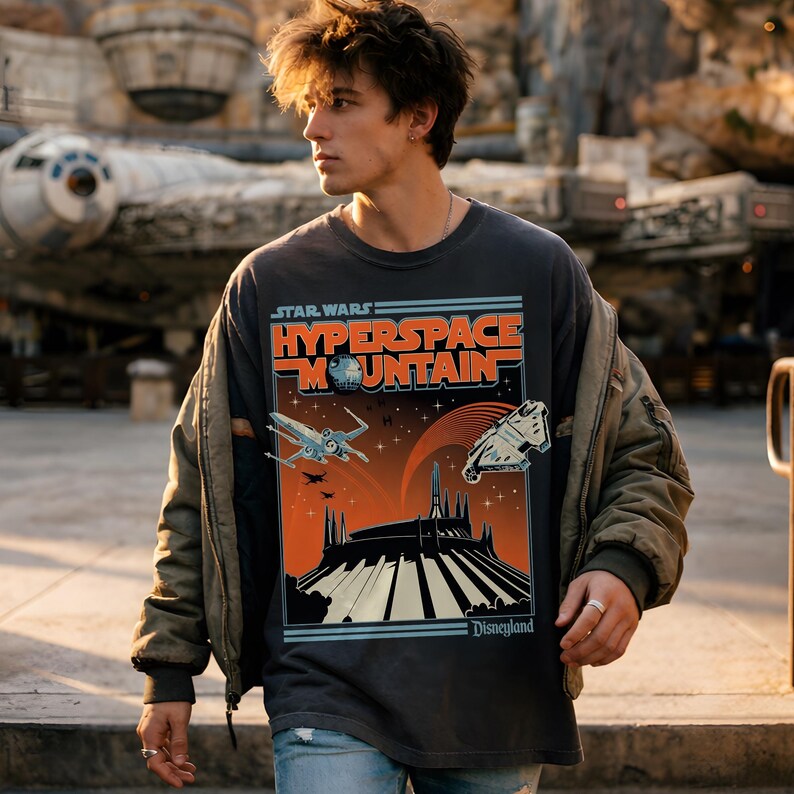 May include: Dark gray Star Wars Hyperspace Mountain t-shirt with a graphic of a spaceship, mountain, and the Disneyland logo. The shirt is worn with a green jacket and blue jeans.