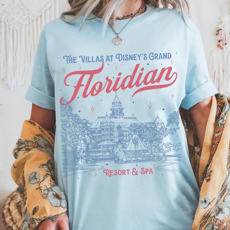May include: Light blue t-shirt with the text "The Villas at Disney's Grand Floridian Resort & Spa" in red and blue. The graphic depicts a detailed illustration of the resort. The shirt has rolled-up sleeves.