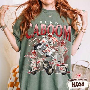 May include: Moss green t-shirt featuring the text "DUKE CABOOM" in red and white, with a graphic of a toy character on a motorcycle. The shirt is labeled "COMFORT COLOR MOSS".