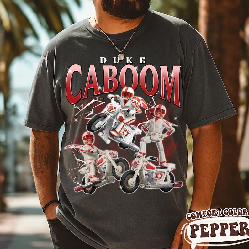May include: Dark gray t-shirt featuring the text "DUKE CABOOM" in red with a white outline. The graphic shows a cartoon character on a motorcycle with red and white accents. The shirt also has the text "COMFORT COLOR PEPPER".