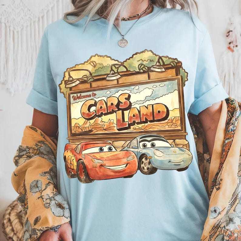 Op de afbeelding: Lichtblauw T-shirt met een Cars Land-afbeelding met Lightning McQueen en Sally Carrera. De afbeelding bevat de tekst "Welcome to Cars Land". Het shirt heeft opgerolde mouwen.