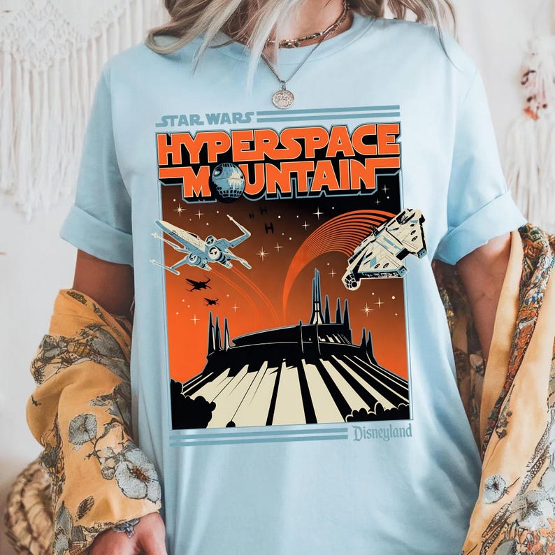 May include: Light blue t-shirt featuring a retro-style graphic for Star Wars: Hyperspace Mountain at Disneyland. The design includes the text "Hyperspace Mountain" and images of the Death Star, X-wing fighters, and the Millennium Falcon.