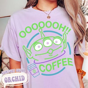 Puede incluir: Una camiseta de color lila claro con un dise&ntilde;o de alien&iacute;gena verde de tres ojos con el texto "OOOOOOH!! COFFEE". Tambi&eacute;n hay un gr&aacute;fico de una taza de caf&eacute;. La camiseta est&aacute; etiquetada como "COMFORT COLOR ORCHID".