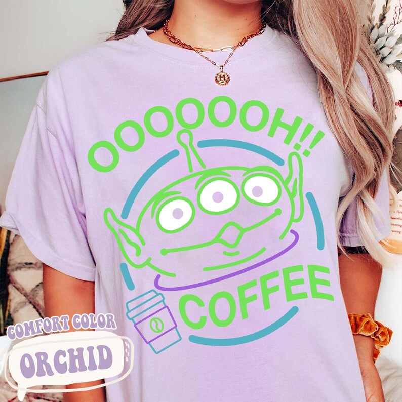 Puede incluir: Una camiseta de color lila claro con un dise&ntilde;o de alien&iacute;gena verde de tres ojos con el texto "OOOOOOH!! COFFEE". Tambi&eacute;n hay un gr&aacute;fico de una taza de caf&eacute;. La camiseta est&aacute; etiquetada como "COMFORT COLOR ORCHID".