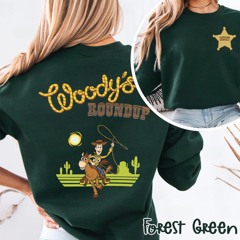 K&ouml;nnte beinhalten: Forstgr&uuml;nes Sweatshirt mit einer Grafik von Woody auf einem Pferd und dem Text "Woody's Roundup". Das Design beinhaltet eine Sonne, Kakteen und das Wort "Sheriff" auf der Vorderseite. Das Sweatshirt hat einen Rundhalsausschnitt.