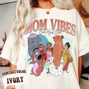 May include: A white t-shirt with a graphic print of cartoon characters from Disney movies. The text "MOM VIBES Cool Moms Club" is printed in pink on the shirt.