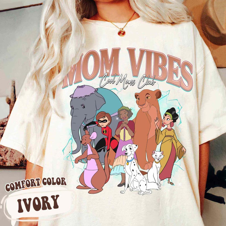 May include: A white t-shirt with a graphic print of cartoon characters from Disney movies. The text "MOM VIBES Cool Moms Club" is printed in pink on the shirt.