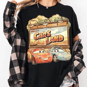 Op de afbeelding: Zwart T-shirt met een Cars Land-afbeelding. Het ontwerp bevat de tekst "Welcome to Cars Land" boven illustraties van Bliksem McQueen en Sally Carrera. Het shirt heeft een relaxte pasvorm.