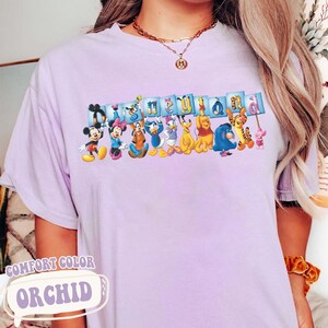 Puede incluir: Camiseta Comfort Colors color orqu&iacute;dea con la palabra "Disneyland" en una pancarta azul con varios personajes de Disney. Mickey Mouse, Minnie Mouse, Pato Donald y Winnie the Pooh se encuentran entre los personajes. La camiseta tambi&eacute;n tiene las palabras "Comfort Color" y "Orchid".