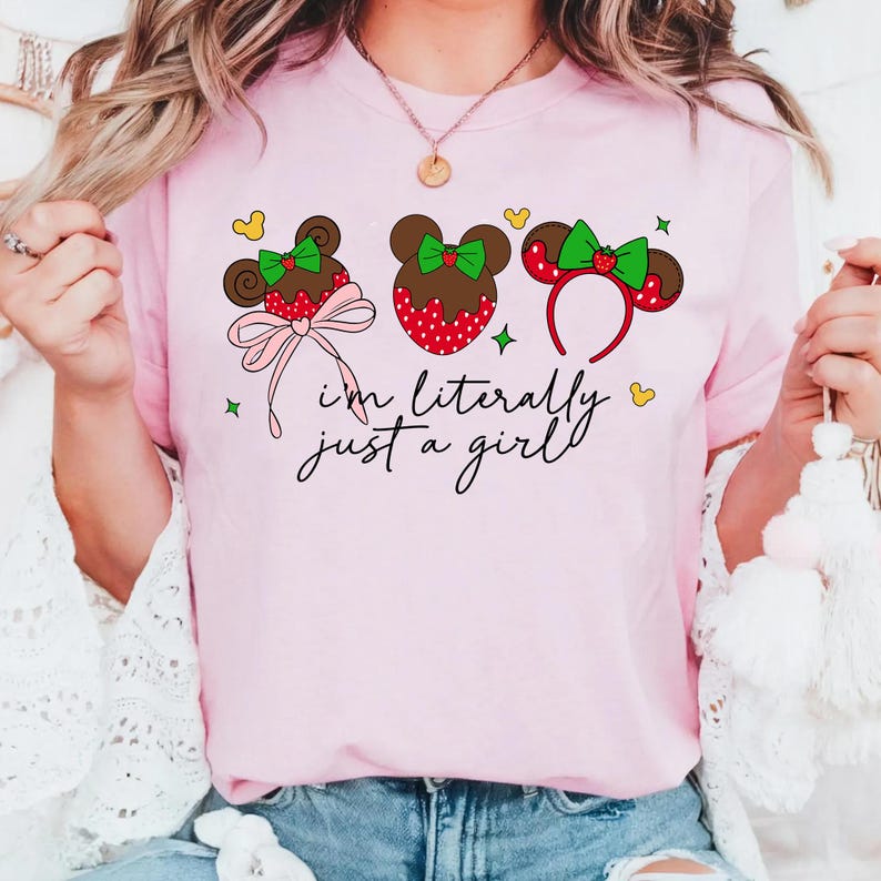 May include: Pink t-shirt featuring three cartoon mouse ear designs with red and white polka dots, green bows, and a strawberry accent. The text "I'm literally just a girl" is written in black script. Yellow hearts and green stars are scattered around the design.