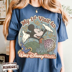 May include: Navy blue t-shirt with a colorful graphic featuring Mickey Mouse, the text "My 1st Visit to Walt Disney World", a wizard hat, a castle, and a geometric sphere. The shirt is labeled "Comfort Color Midnight".