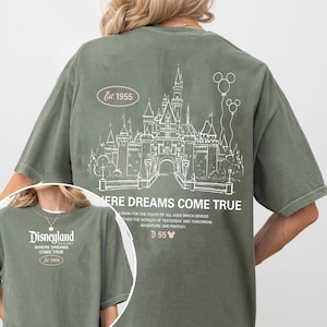 May include: Olive green t-shirt featuring a white line drawing of a castle and balloons. The text "Where Dreams Come True" and "Disneyland" are also visible. The shirt includes the text "Est. 1955" and "D55".