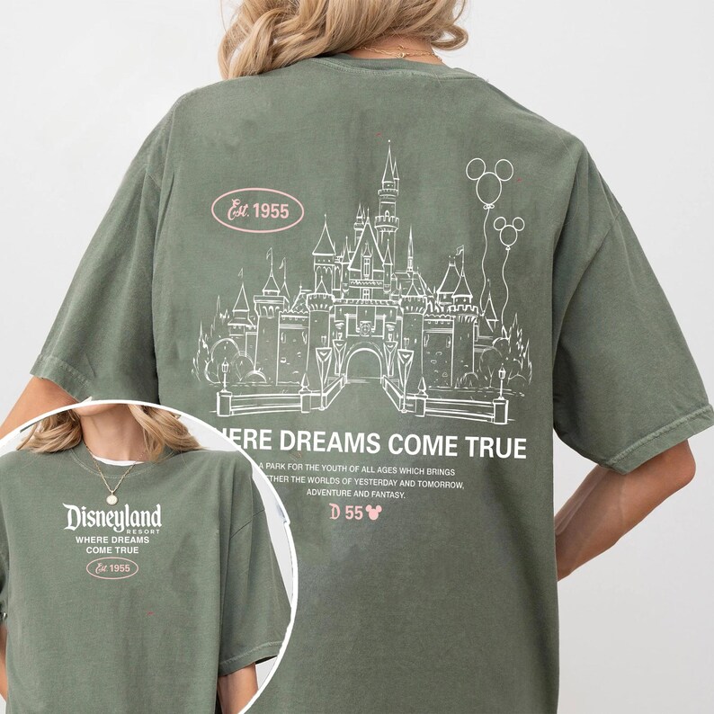 May include: Olive green t-shirt featuring a white line drawing of a castle and balloons. The text "Where Dreams Come True" and "Disneyland" are also visible. The shirt includes the text "Est. 1955" and "D55".
