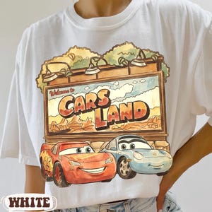 Op de afbeelding: Wit T-shirt met een kleurrijke afbeelding van Cars Land met Bliksem McQueen en Sally Carrera. Het ontwerp bevat de tekst "Welcome to Cars Land" en het nummer "95" op Bliksem McQueen. Een leuk ontwerp in cartoonstijl.