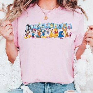 Puede incluir: Camiseta rosa claro con un dise&ntilde;o colorido de personajes de Disney, incluyendo Mickey Mouse, Minnie Mouse, Donald y Winnie the Pooh. La palabra "Disneyland" est&aacute; escrita en letras azules sobre los personajes.