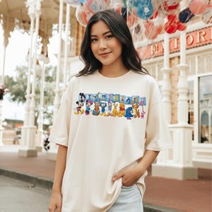 Puede incluir: Camiseta color crema con un gr&aacute;fico colorido de personajes de Disney, incluyendo Mickey Mouse, Minnie Mouse y el Pato Donald. La palabra "Disneyland" se muestra encima de los personajes en letras azules. La camiseta la lleva una persona.