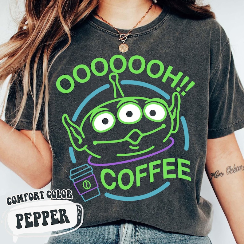 Puede incluir: Camiseta gris oscuro con un gr&aacute;fico de un alien&iacute;gena verde con tres ojos, el texto "OOOOOOH!! COFFEE" y una taza de caf&eacute;. La camiseta tambi&eacute;n tiene el texto "COMFORT COLOR PEPPER".