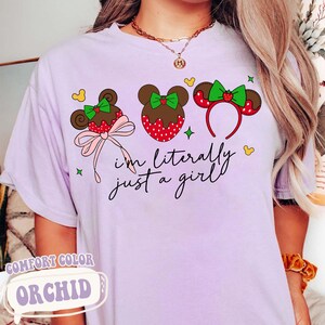 May include: A light purple t-shirt with a graphic of three mouse ear designs with bows and strawberries. The text "I'm literally just a girl" is written in black script. The shirt is labeled "Comfort Color Orchid".
