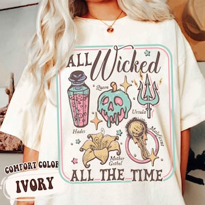May include: Ivory-colored t-shirt with a graphic design featuring the text "ALL Wicked ALL THE TIME." The design includes illustrations of a poison bottle, a skull, a trident, a flower, and a magic mirror. The shirt is labeled "COMFORT COLOR."