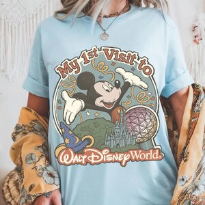 May include: Light blue t-shirt featuring a colorful graphic of Mickey Mouse with the text "My 1st Visit to Walt Disney World." The design includes a wizard hat, a castle, and a geometric sphere. The shirt has short sleeves.