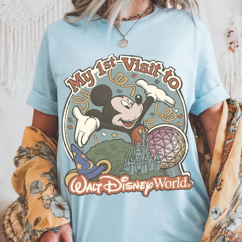 May include: Light blue t-shirt featuring a colorful graphic of Mickey Mouse with the text "My 1st Visit to Walt Disney World." The design includes a wizard hat, a castle, and a geometric sphere. The shirt has short sleeves.