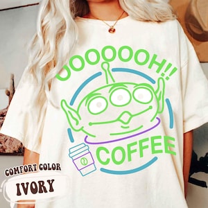 Puede incluir: Camiseta marfil Comfort Colors con un gr&aacute;fico de alien&iacute;gena verde y el texto "OOOOH!! COFFEE". El alien&iacute;gena tiene tres ojos y orejas grandes. Un contorno azul y morado rodea al alien&iacute;gena y la taza de caf&eacute;.