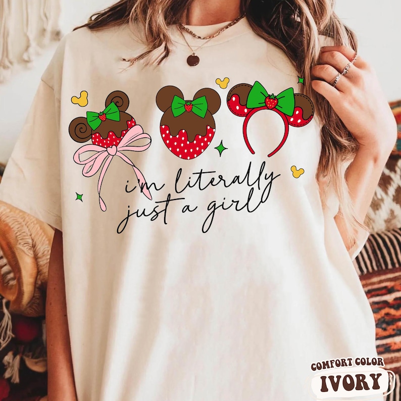 May include: Ivory-colored t-shirt featuring three Mickey Mouse-inspired designs with a strawberry theme. The text "I'm literally just a girl" is written in cursive. The shirt has a Comfort Color label.