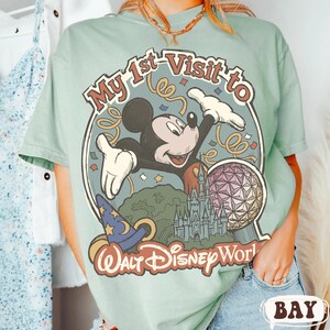 May include: A light green t-shirt featuring a graphic of Mickey Mouse with the text "My 1st Visit to Walt Disney World." The design includes a castle, a sphere, and a wizard's hat.