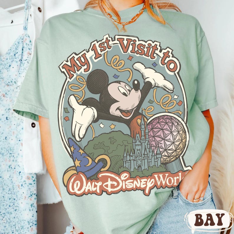 May include: A light green t-shirt featuring a graphic of Mickey Mouse with the text "My 1st Visit to Walt Disney World." The design includes a castle, a sphere, and a wizard's hat.