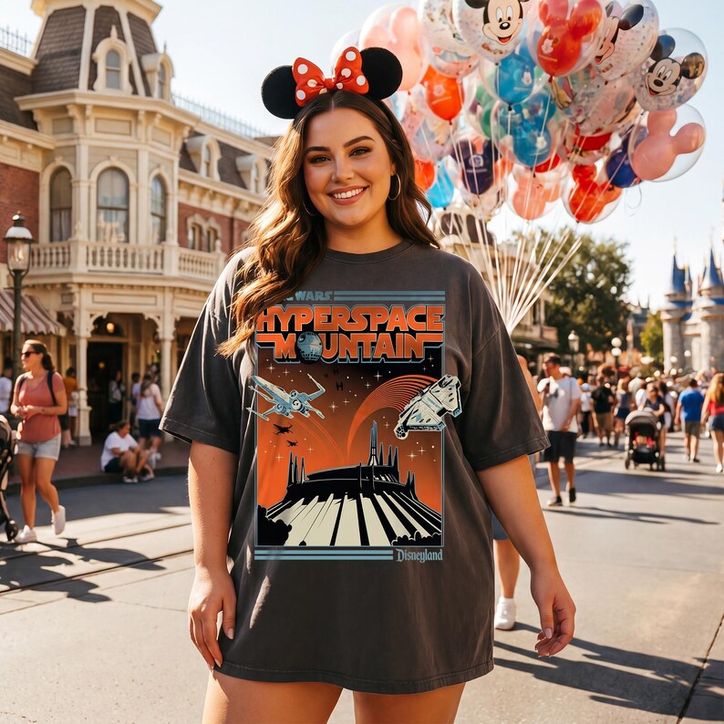 May include: A dark gray t-shirt featuring a graphic for the Star Wars: Hyperspace Mountain ride at Disneyland. The design includes spaceships, a mountain silhouette, and the Disneyland logo. The person is wearing Minnie Mouse ears and holding balloons.